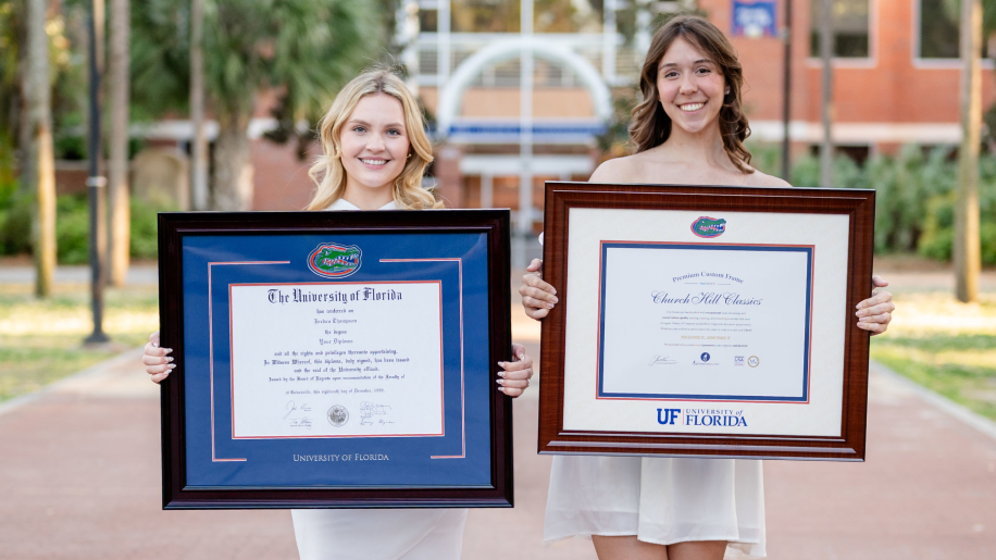 Two University of Florida graduates in white dresses standing on campus and proudly holding framed diplomas from Church Hill Classics