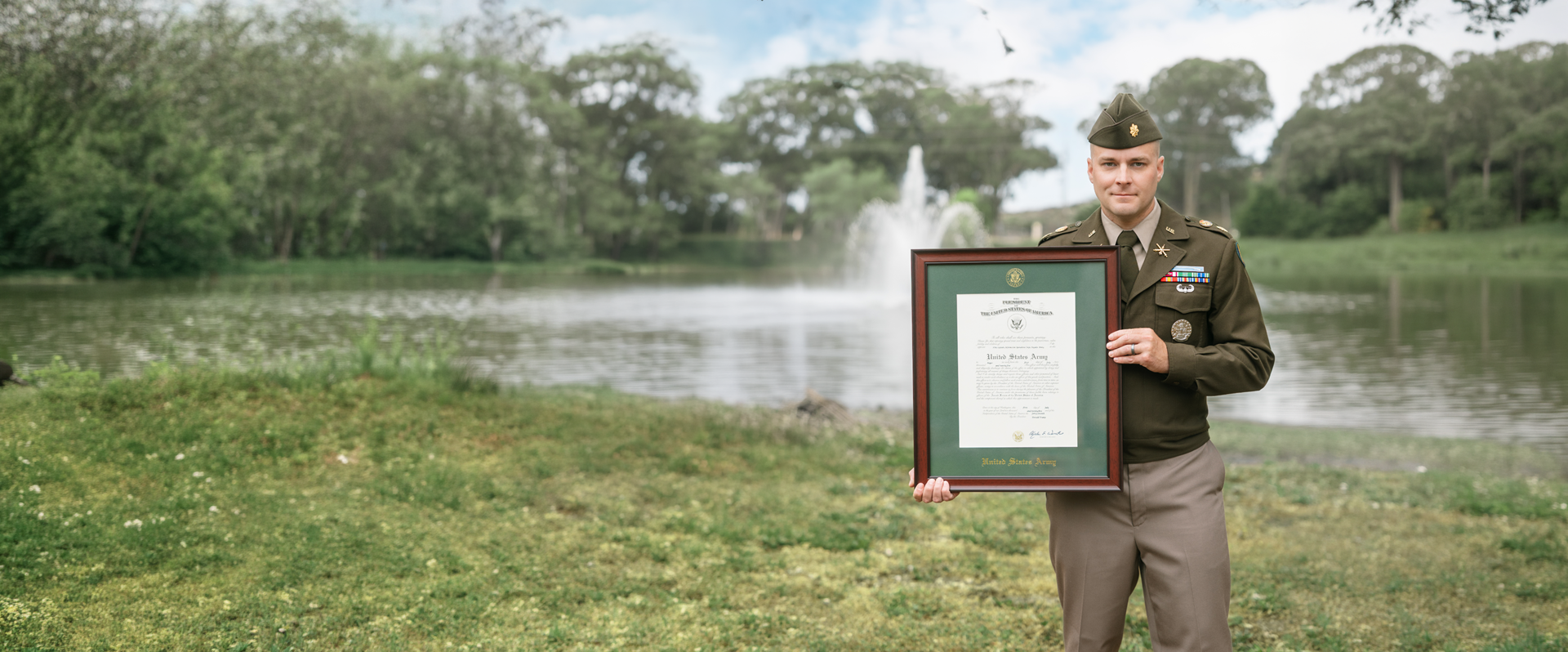 U.S. Army service member standing outdoors by a lake, holding a framed military commission certificate with green matting and a dark wood frame.