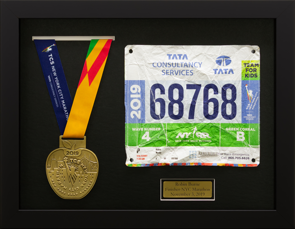 Framed New York City Marathon medal and race bib from 2019, mounted on a black background with a commemorative plaque.