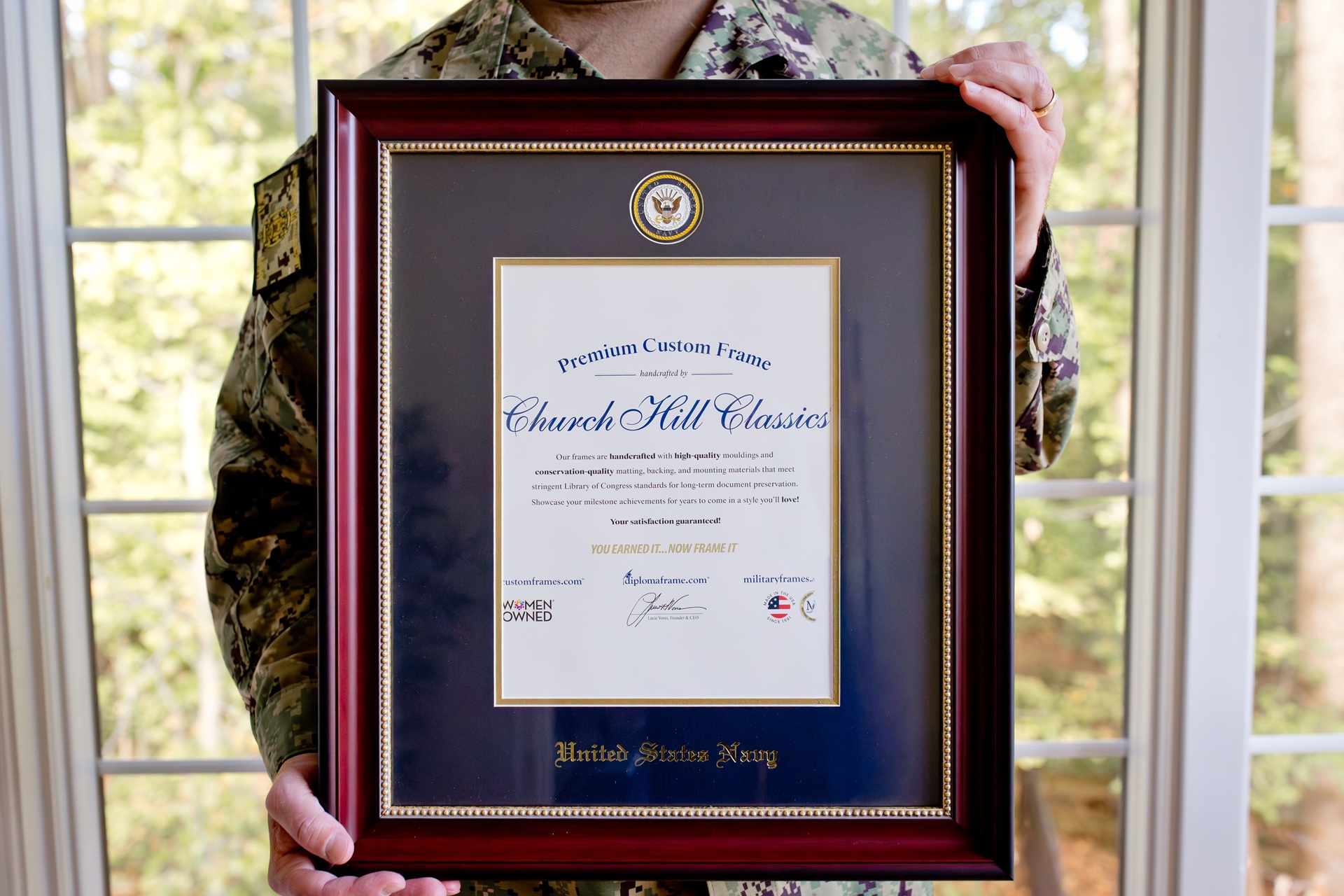 Person in a U.S. Navy camouflage uniform holding a framed certificate display with navy blue matting, gold accents, and a United States Navy emblem above the document.