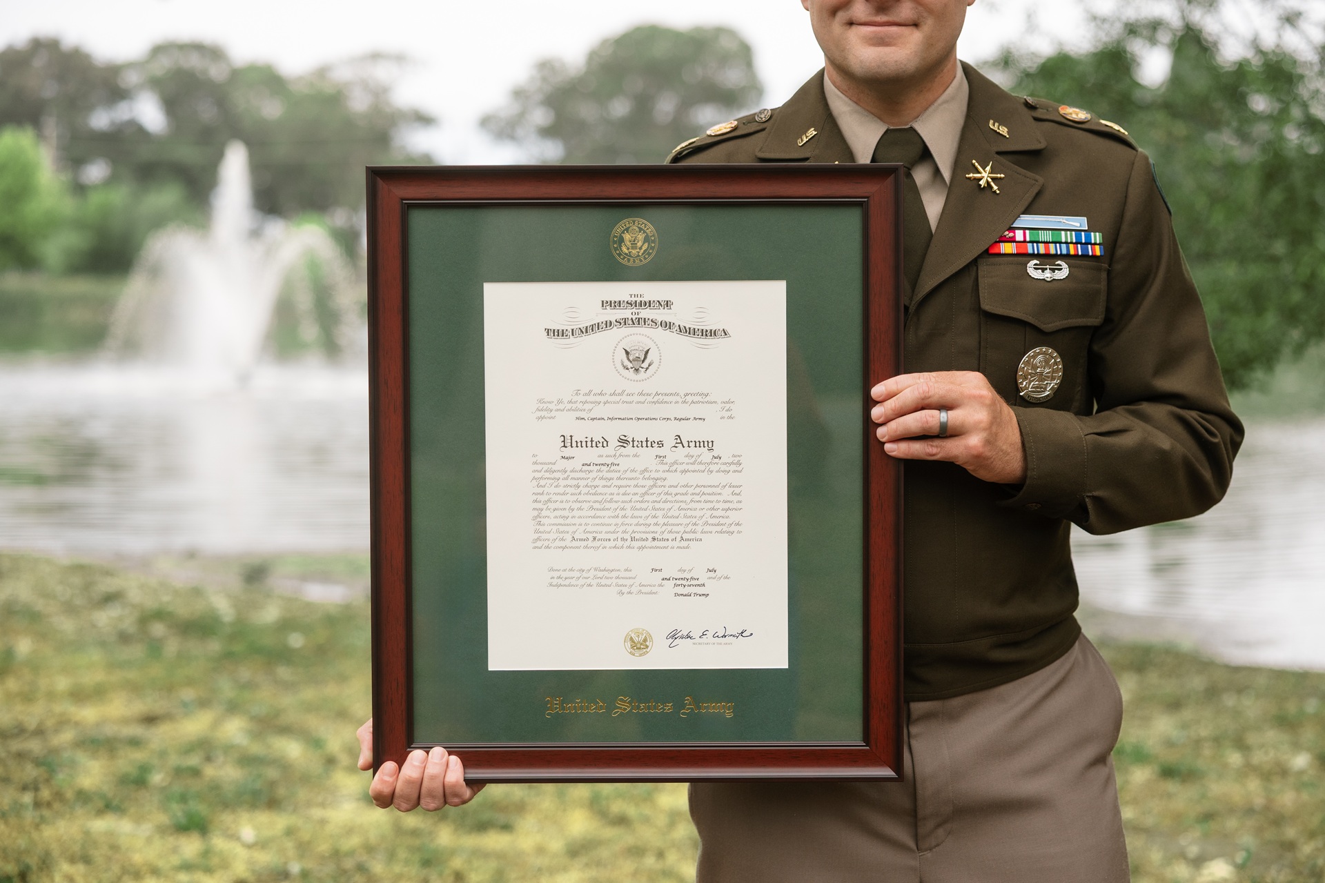 U.S. Army service member standing outdoors near a lake, holding a framed military commission certificate with green matting, a wood frame, and the United States Army seal.