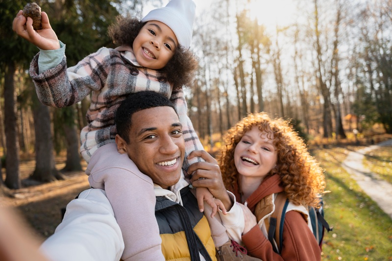 Smiling family taking a selfie outdoors in a wooded park, with a child sitting on an adult’s shoulders during golden hour.
