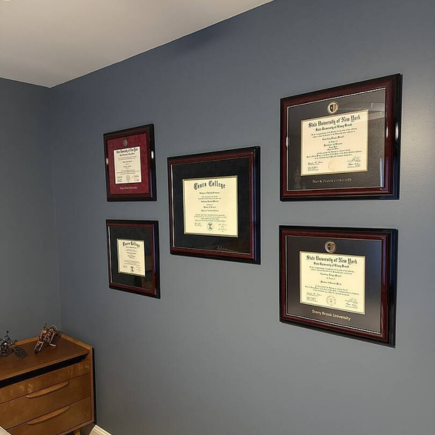 Gallery wall featuring five framed diplomas from various institutions, arranged neatly on a slate-blue wall above a mid-century modern dresser.