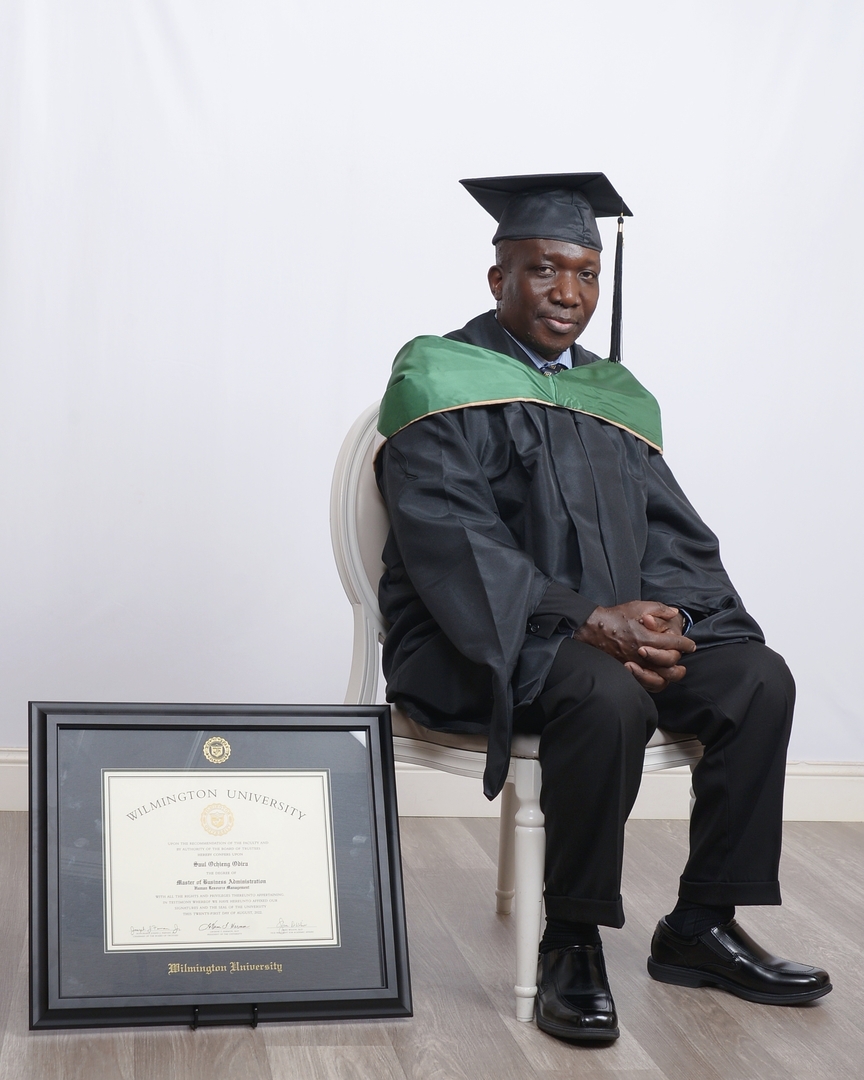 man in graduation regalia sitting next to wilmington university frame