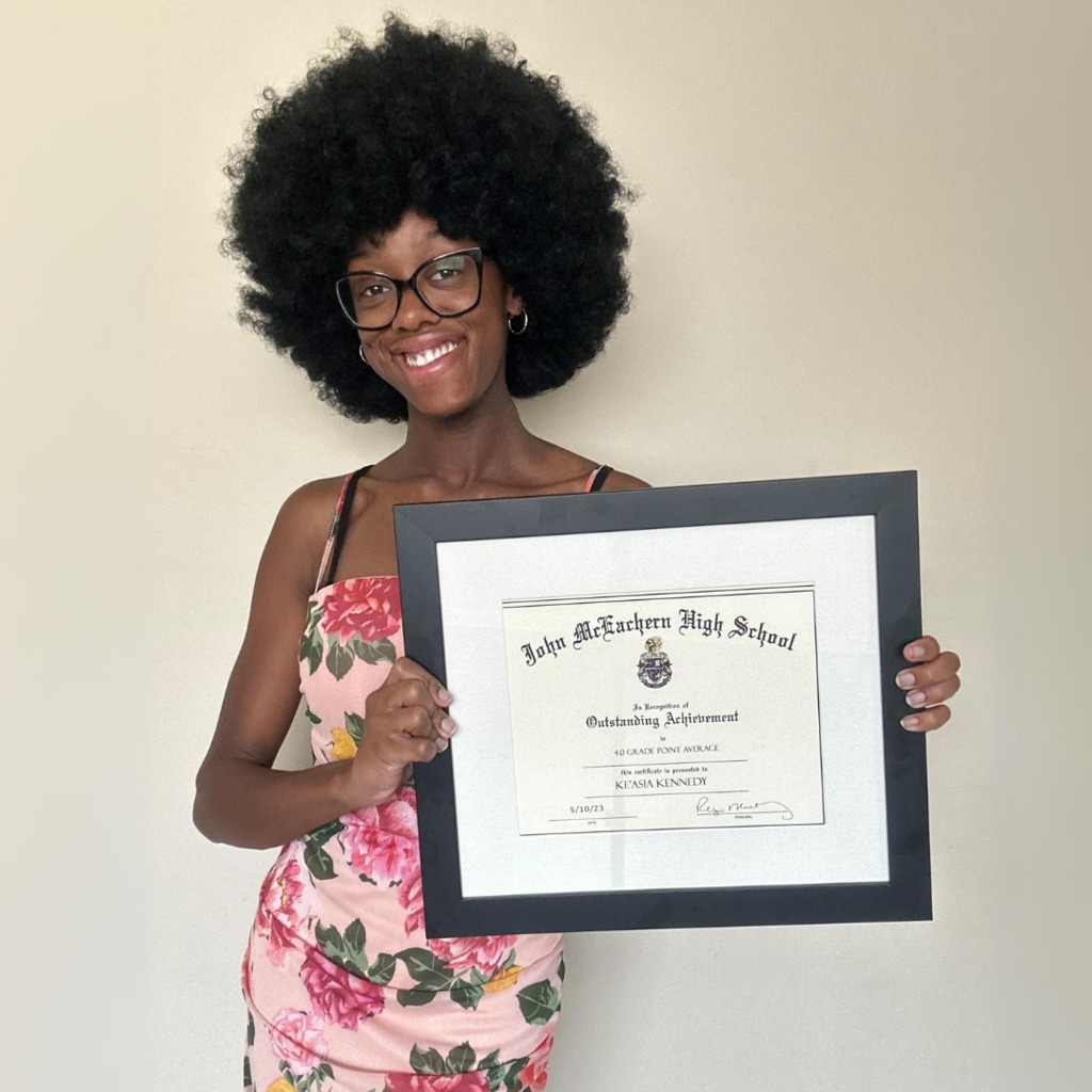 smiling teenager in floral dress holding her framed high school diploma