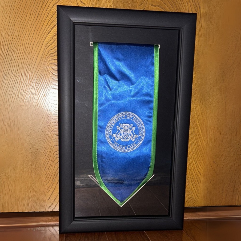 closeup of University of Houston Clear Lake graduation stole frame leaning against wood grain wall