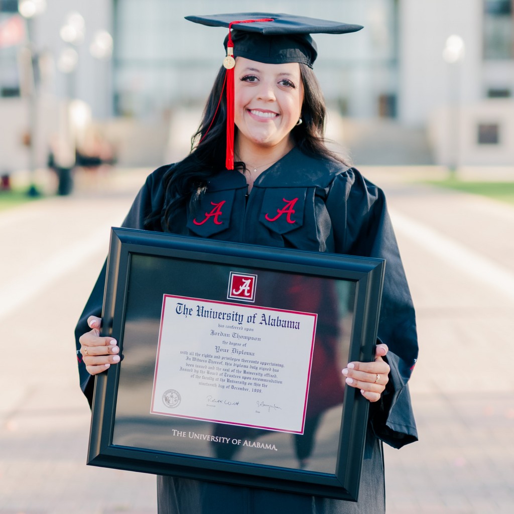 Graduate on campus holding University of Alabama diploma frame