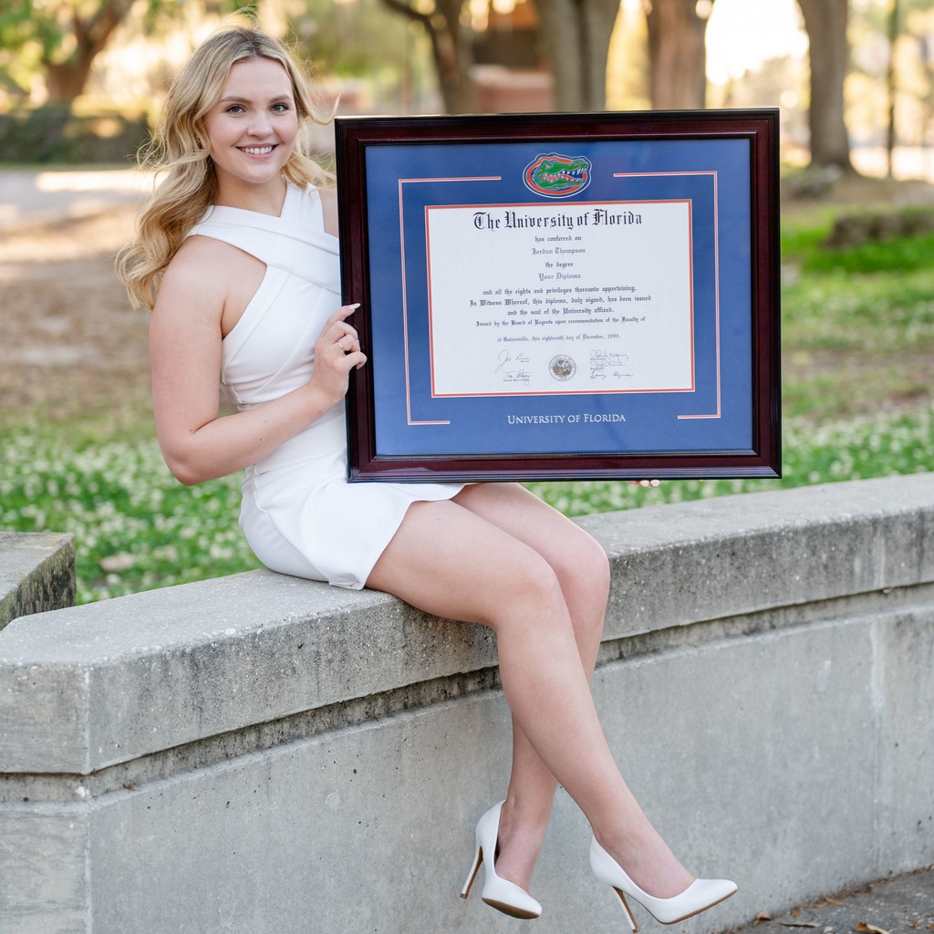 smiling girl in white dress sitting on ledge outdoors holding University of Florida Gators diploma frame