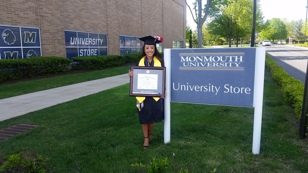 female graduate wearing cap and gown holding a Monmouth University diploma frame outside